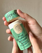 A person holds an open tube of Good Flower Farm Pine Cedarwood Deodorant Plastic-Free Tube, labeled as aluminum- and baking soda-free, in biodegradable packaging against a plain background.