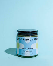 A glass jar labeled Good Flower Farm Good Baby Chapped Cheeks Diaper Balm with a floral design and black lid sits on a blue surface. The 99g (3.5oz) balm soothes delicate baby skin against a light blue background.