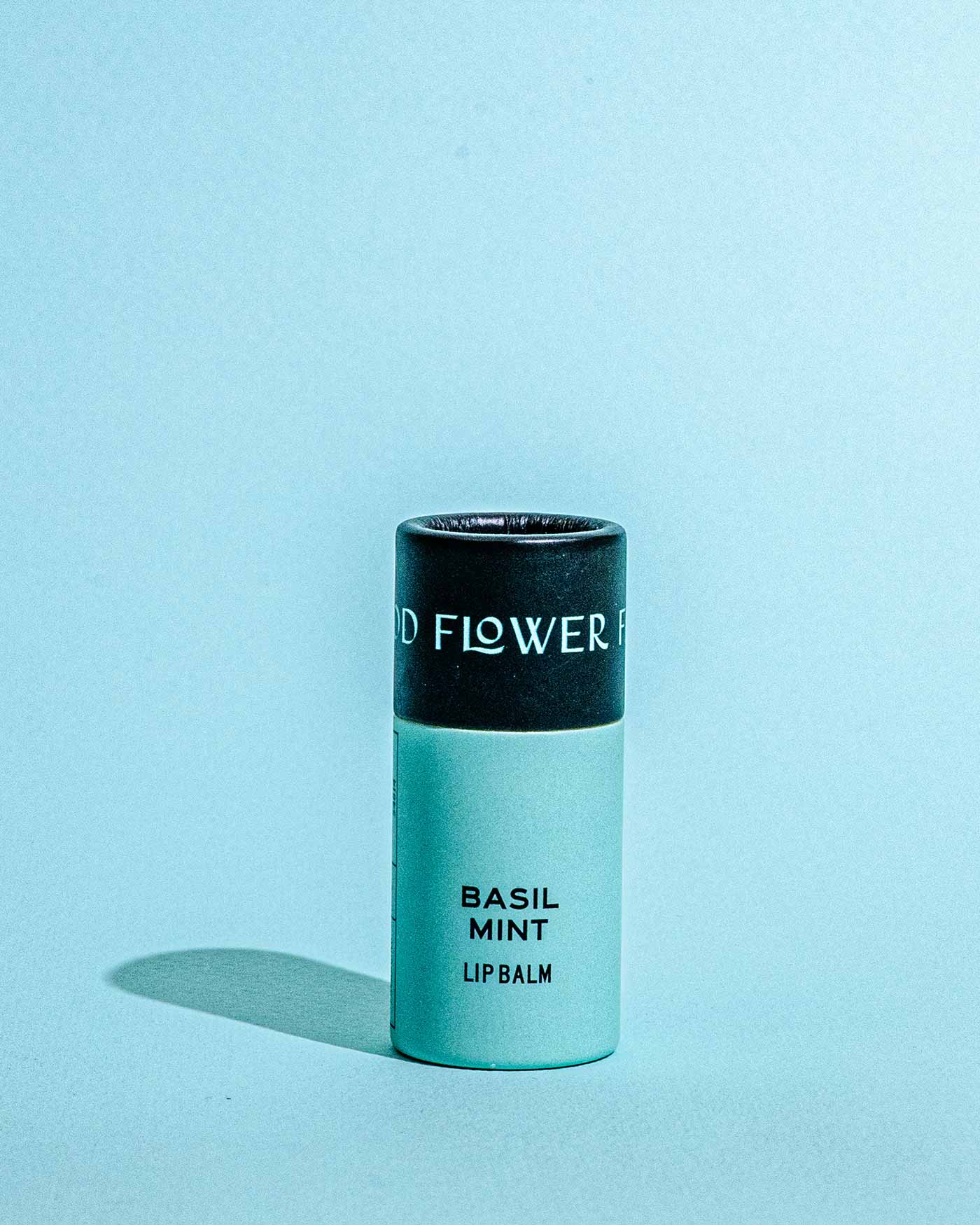 A cylindrical tube of Good Flower Farms TESTER — Basil Mint Lip Organic Lip Balm stands upright against a light blue background, with a black cap and light green body. The label reads Basil Mint Lip Balm. A soft shadow falls to the left.
