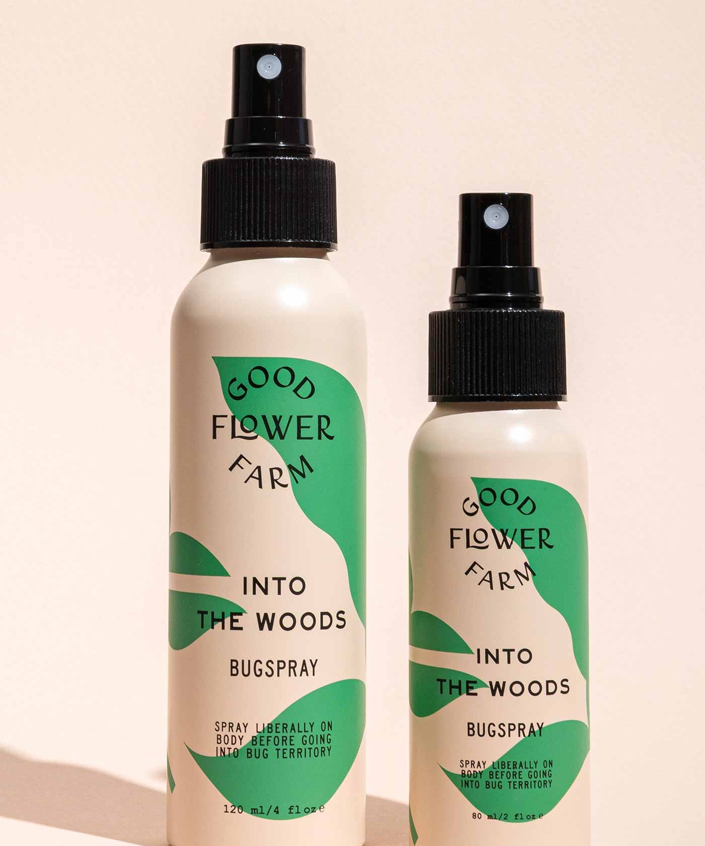 Two Good Flower Farm Into the Woods Natural Bugsprays, one large and one small, with green and cream labels, stand side by side on a beige background—the smaller is crafted with essential oils.