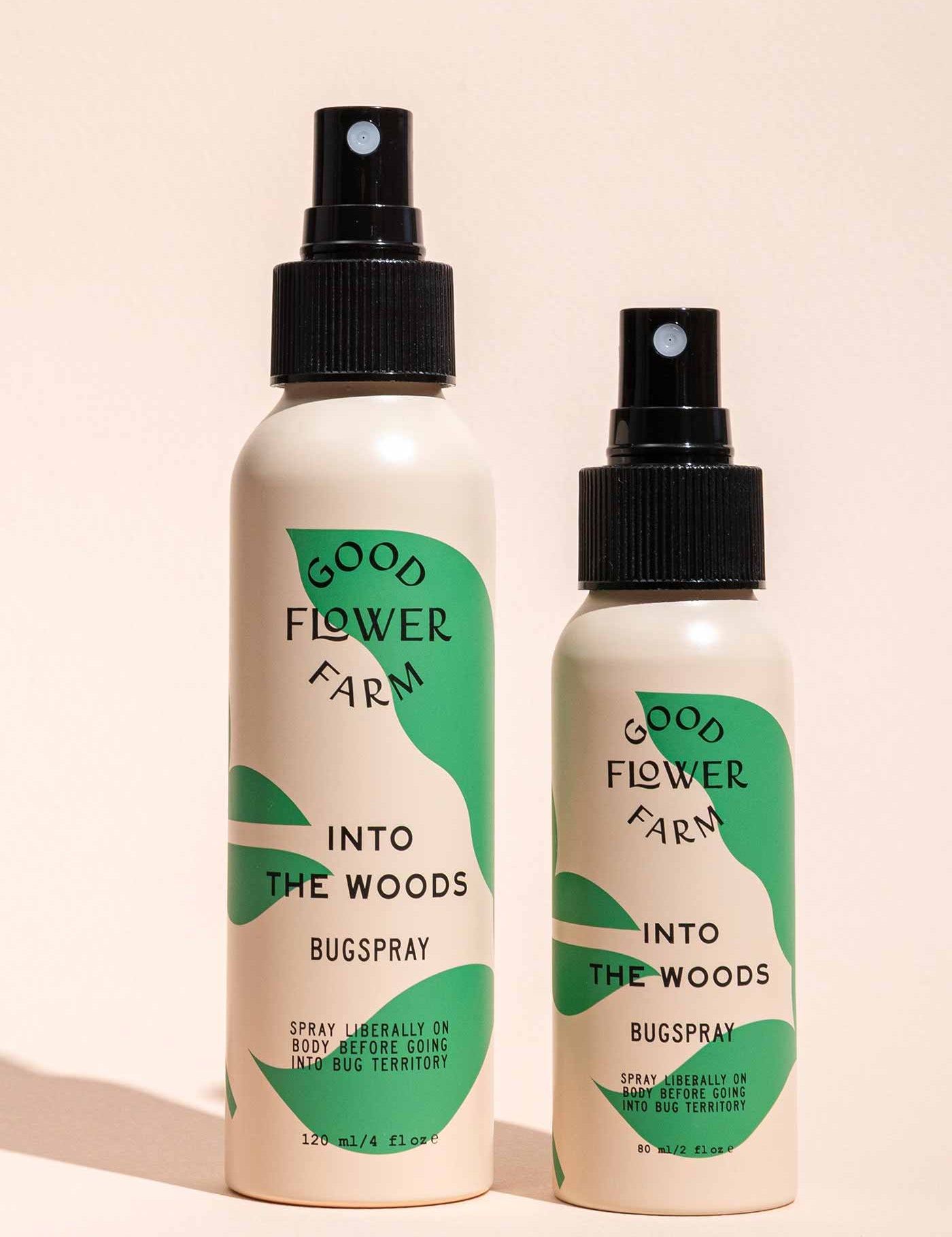Two Good Flower Farm Into the Woods Natural Bugsprays, one large and one small, with green and cream labels, stand side by side on a beige background—the smaller is crafted with essential oils.