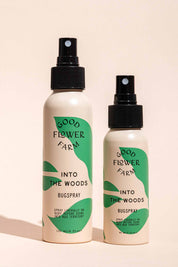 Two Good Flower Farm Into the Woods Natural Bugsprays, one large and one small, with green and cream labels, stand side by side on a beige background—the smaller is crafted with essential oils.
