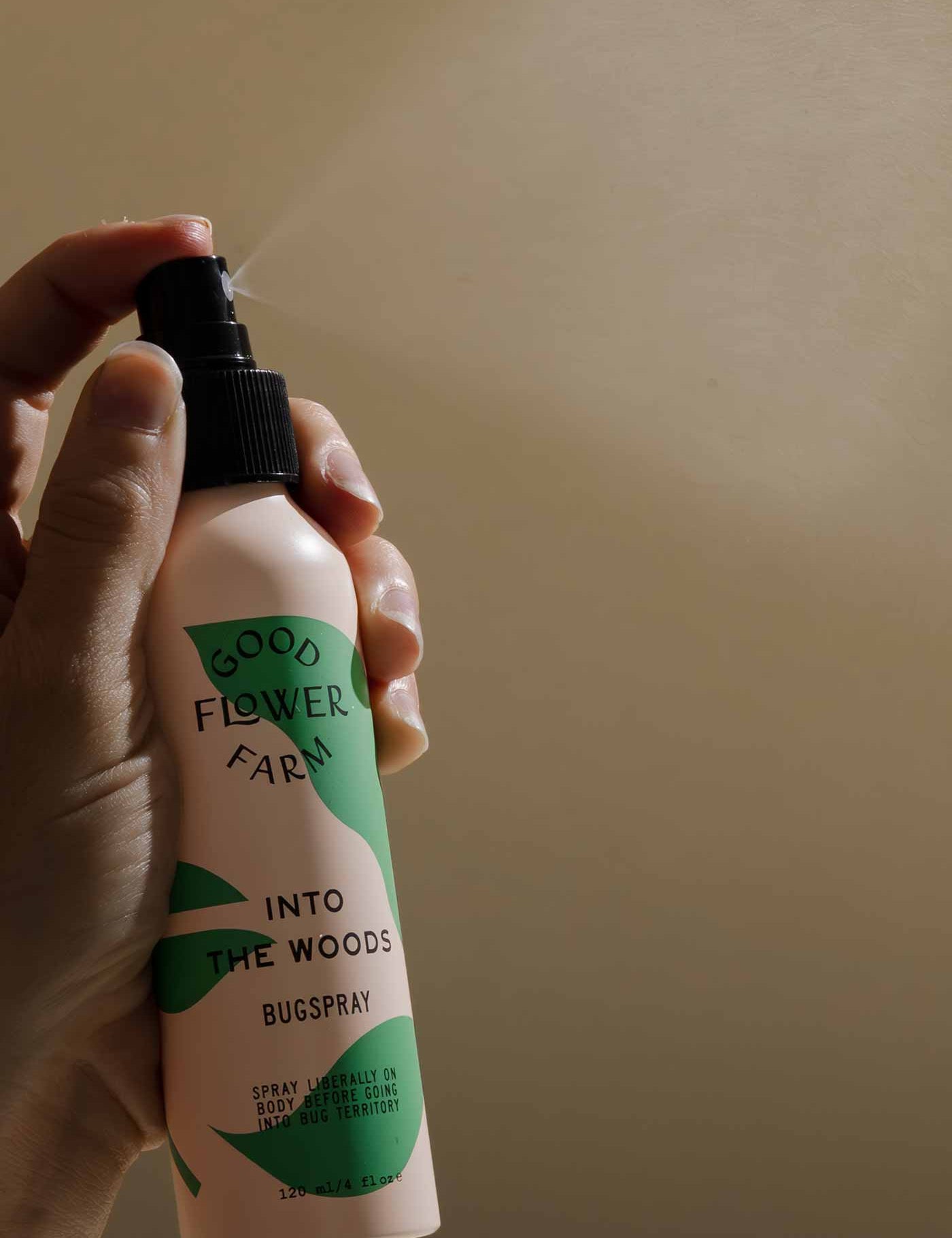 A hand holds a bottle of Good Flower Farm Into the Woods Natural Bugspray, an organic insect repellent with essential oils, set against a softly lit beige background.