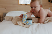 A baby on a bed reaches for the Good Flower Farm Good Baby Gift Set—featuring gentle skincare products—placed on a striped cloth in a cozy, wood-paneled room.