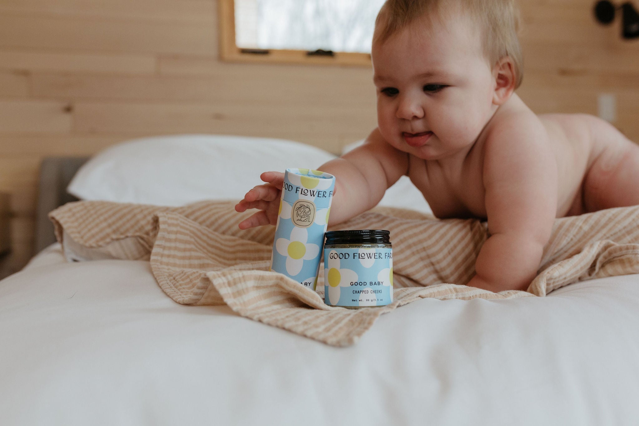 A baby on a bed reaches for the Good Flower Farm Good Baby Gift Set—featuring gentle skincare products—placed on a striped cloth in a cozy, wood-paneled room.