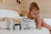 A baby on a bed reaches for the Good Flower Farm Good Baby Gift Set, which includes botanical skincare pouches, herbal baby powder in a jar, and diaper balm in a tube with blue and yellow floral designs.