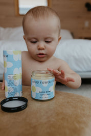 A baby indoors reaches for an open Good Flower Farm Good Baby Gift Set balm on the table, with its packaging and lid nearby; the softly blurred background highlights the botanical-themed product.