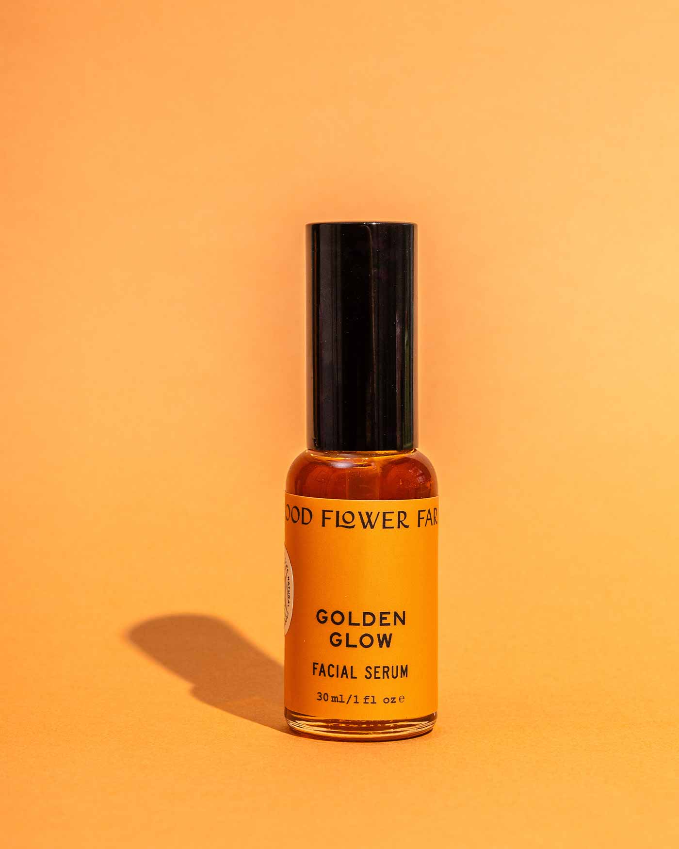 A small amber bottle labeled Golden Glow Facial Serum by Good Flower Farm stands upright with a black cap against an orange background, offering skin hydration and a natural glow.