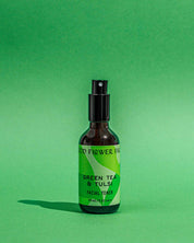 A brown bottle of Good Flower Farm Green Tea & Tulsi Facial Toner with a black spray cap sits against a green background. The green label displays the product name and highlights soothing ingredients: green tea and tulsi.