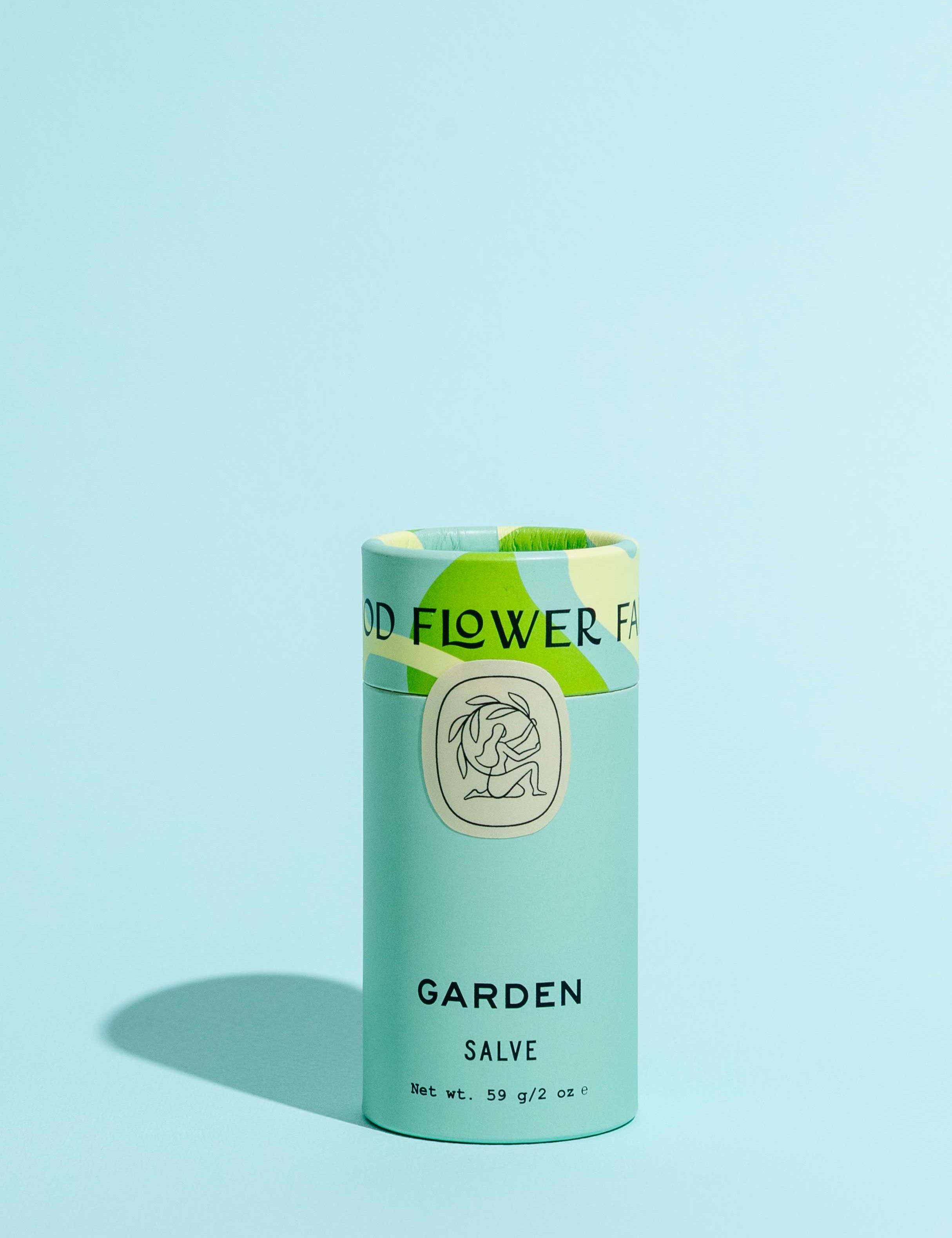 A cylindrical Garden Salve by Good Flower Farm stands upright on a light blue surface with a matching background, featuring light blue packaging, green accents, and an illustrated logo.
