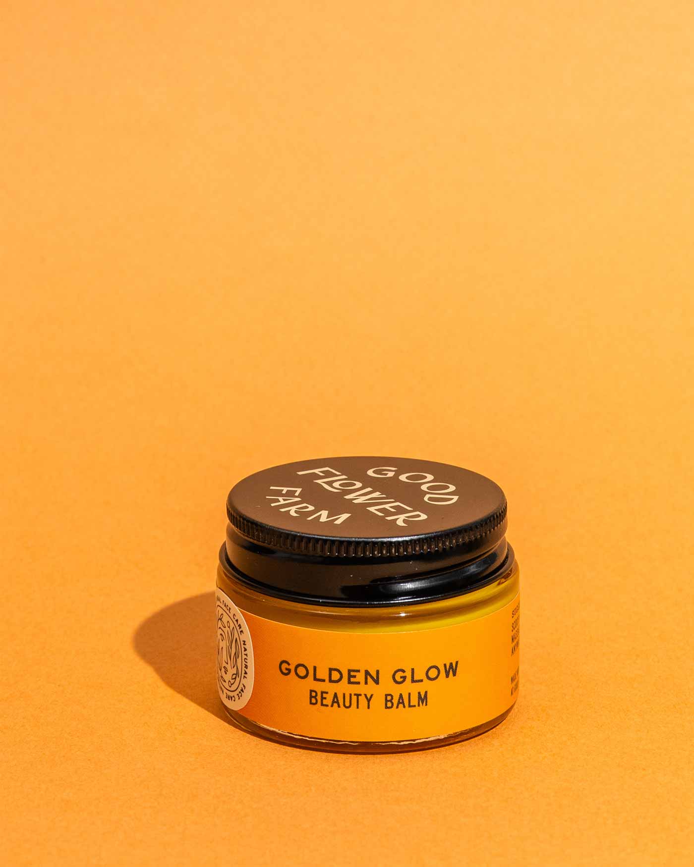 A small amber jar with a black lid labeled Good Flower Farm, featuring the Golden Glow Beauty Balm, sits against a bright orange background—your go-to skin repair balm for a radiant glow.