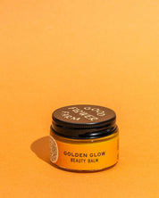 A small amber jar with a black lid labeled Good Flower Farm, featuring the Golden Glow Beauty Balm, sits against a bright orange background—your go-to skin repair balm for a radiant glow.