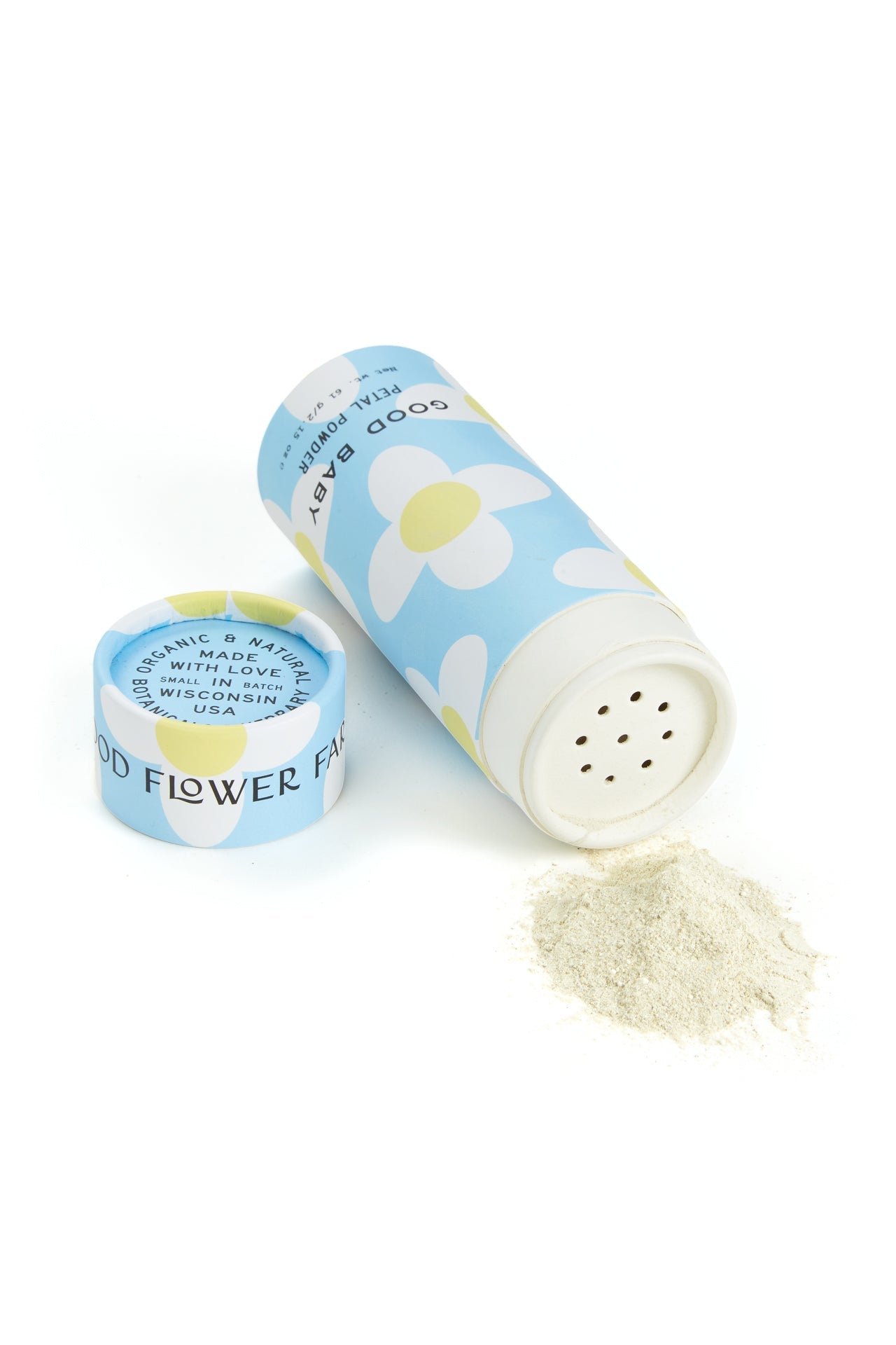 A cylindrical blue container from Good Flower Farm’s Good Baby Gift Set, filled with herbal baby powder, lies open as light-colored powder spills onto a white surface. The cap sits beside it and white and yellow flowers decorate the container.