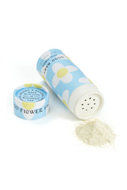 A cylindrical blue container from Good Flower Farm’s Good Baby Gift Set, filled with herbal baby powder, lies open as light-colored powder spills onto a white surface. The cap sits beside it and white and yellow flowers decorate the container.