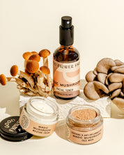 The Good Flower Farm Magic Mushroom Skincare Gift Set, featuring a facial toner, moisturizer, and face mask rich in skin nutrients, is displayed with fresh mushrooms on a beige background.