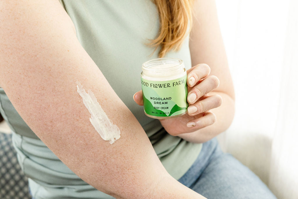 A person applies Good Flower Farm Woodland Dream Organic Body Cream from a jar to their upper arm, leaving a visible streak. They wear a light green sleeveless top and blue jeans.