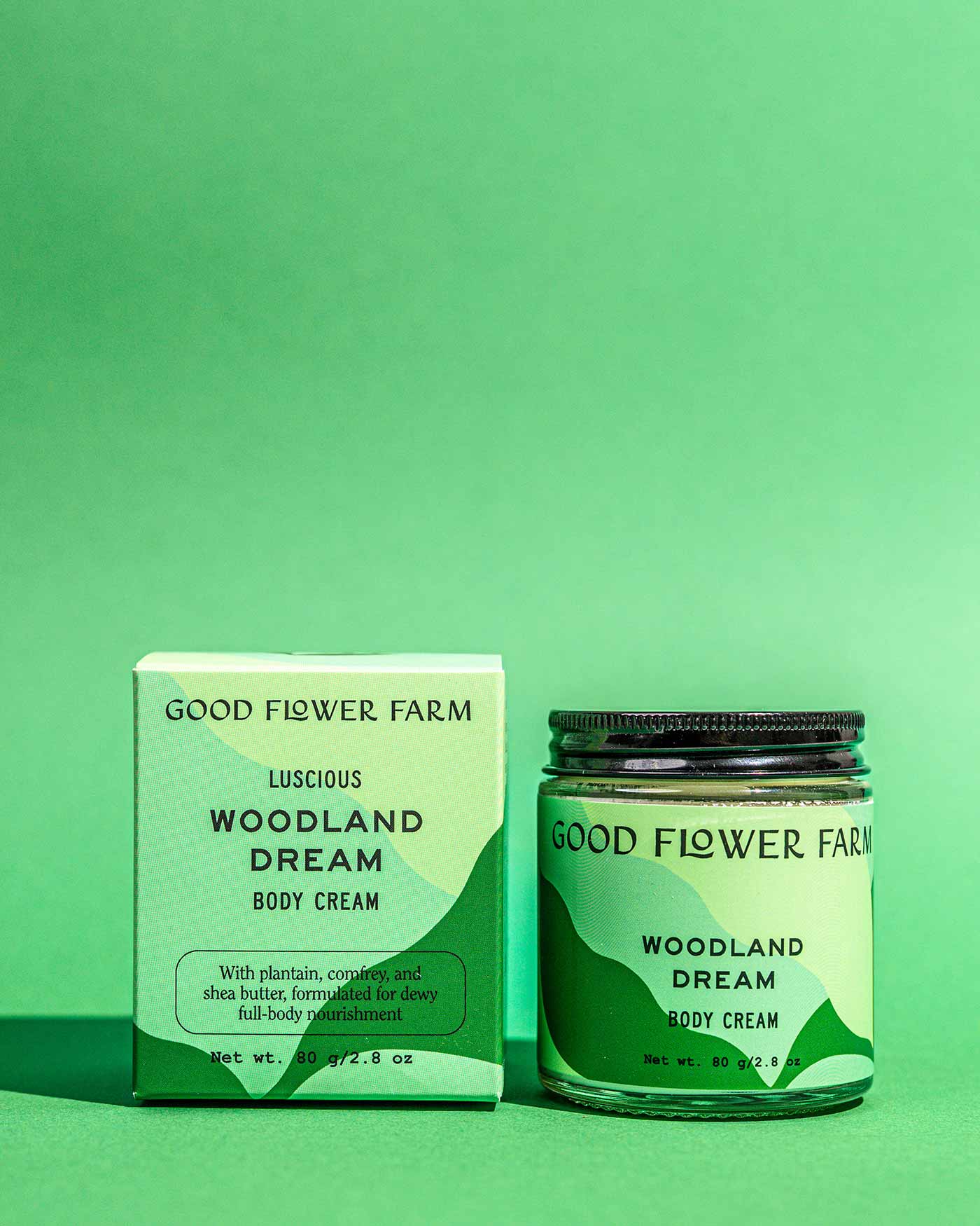 A jar and box of Good Flower Farm Woodland Dream Organic Body Cream are shown on a green background, featuring minimal packaging that highlights botanical ingredients in this shea butter moisturizer.