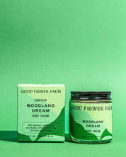 A jar and box of Good Flower Farm Woodland Dream Organic Body Cream are shown on a green background, featuring minimal packaging that highlights botanical ingredients in this shea butter moisturizer.