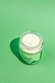 An open glass jar of TESTER — Woodland Dream Organic Body Cream by Good Flower Farm sits on a green background, revealing a swirl of shea butter and Douglas Fir hydrosol-infused cream inside.