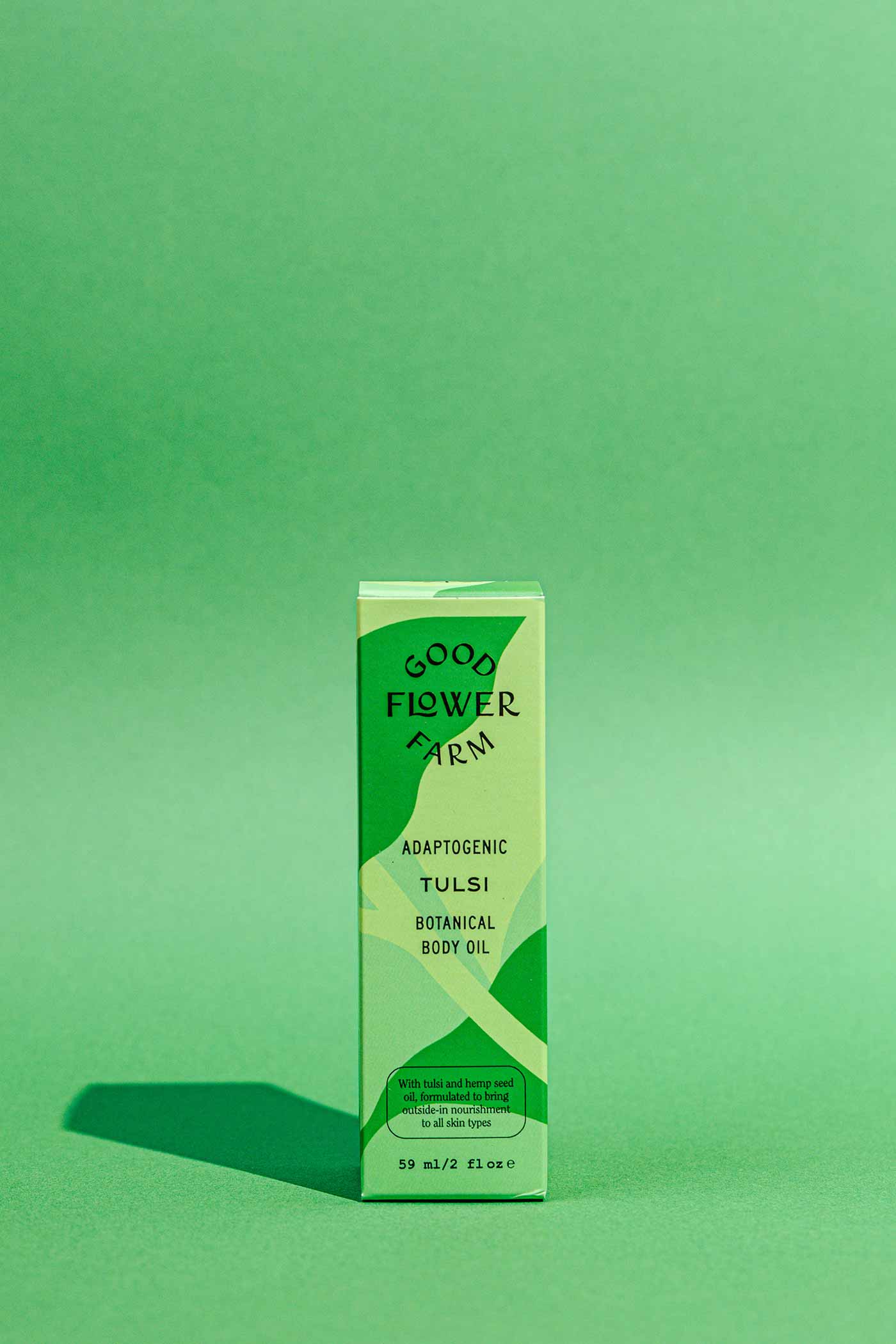 A box of Good Flower Farm Tulsi Botanical Body Oil stands upright on a green surface, with a matching green background and light green geometric patterns featured on the packaging.