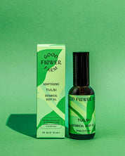 A bottle of Good Flower Farm Tulsi Botanical Body Oil rests beside its green box, both displaying matching green designs and text, set against a green background.