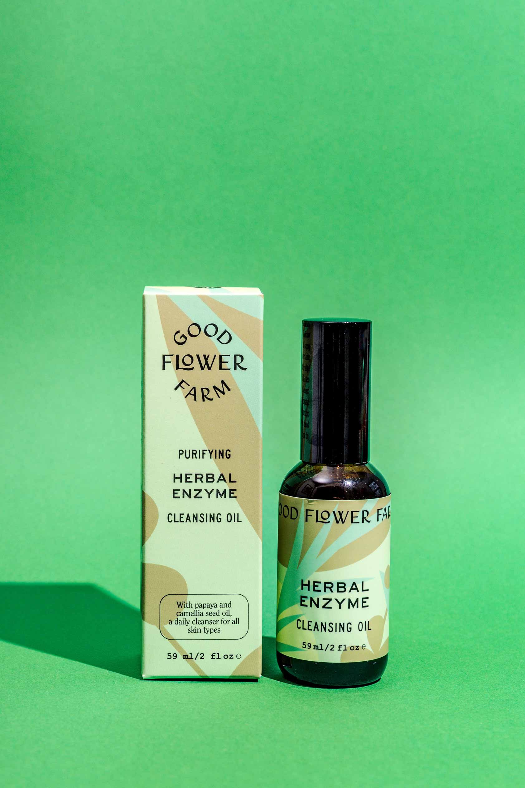 A bottle of Good Flower Farms Herbal Enzyme Cleansing Oil stands beside its matching box, both with green and beige labels, on a solid green background.