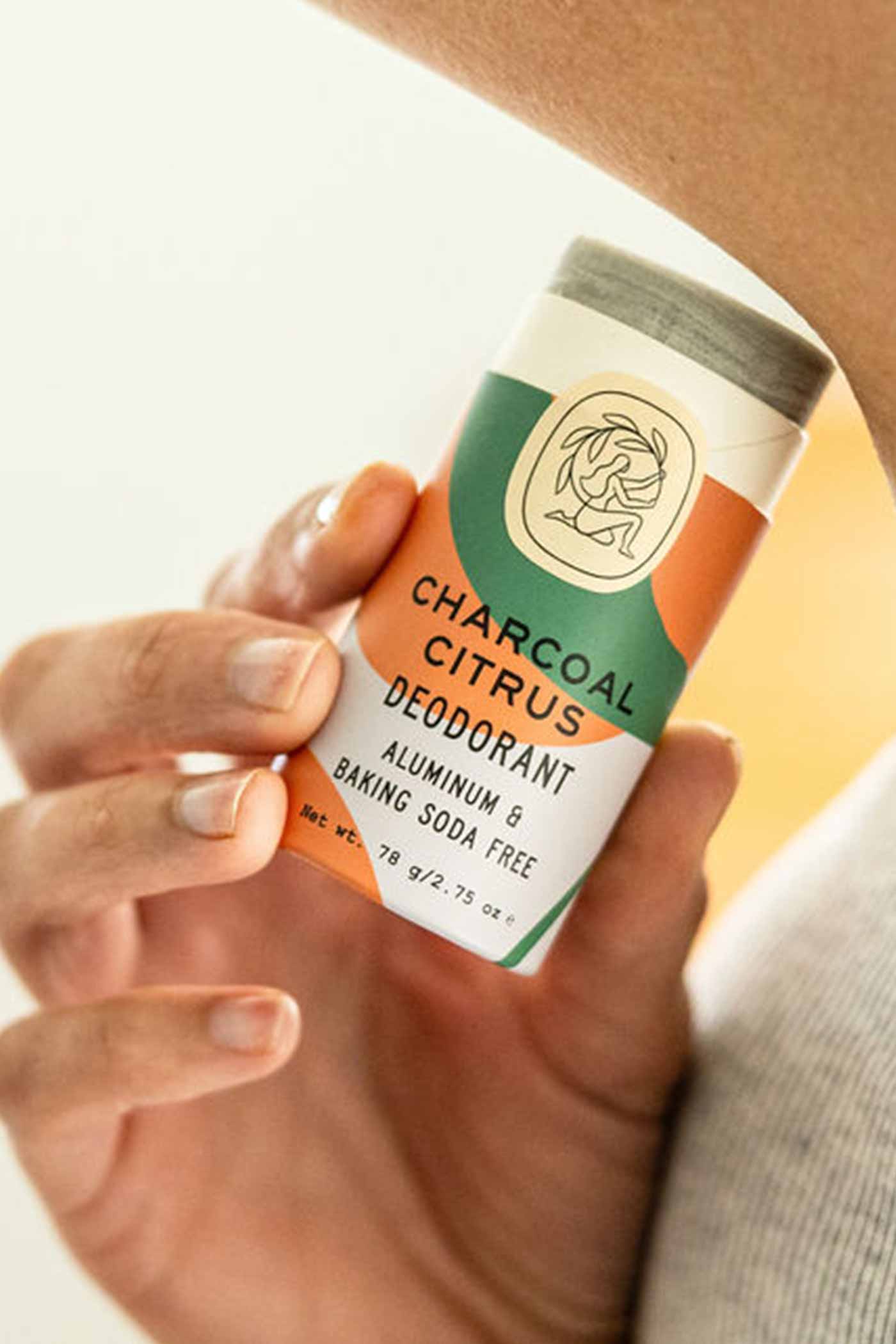 A hand holds a Good Flower Farm Charcoal Citrus Deodorant Plastic-Free Tube, made with essential oils and free from aluminum and baking soda, featuring a vibrant orange, green, and cream design.