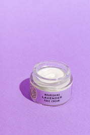 A glass jar of Good Flower Farm’s Lavender Face Cream sits open on a purple surface, revealing the creamy white, calendula-infused moisturizer inside.