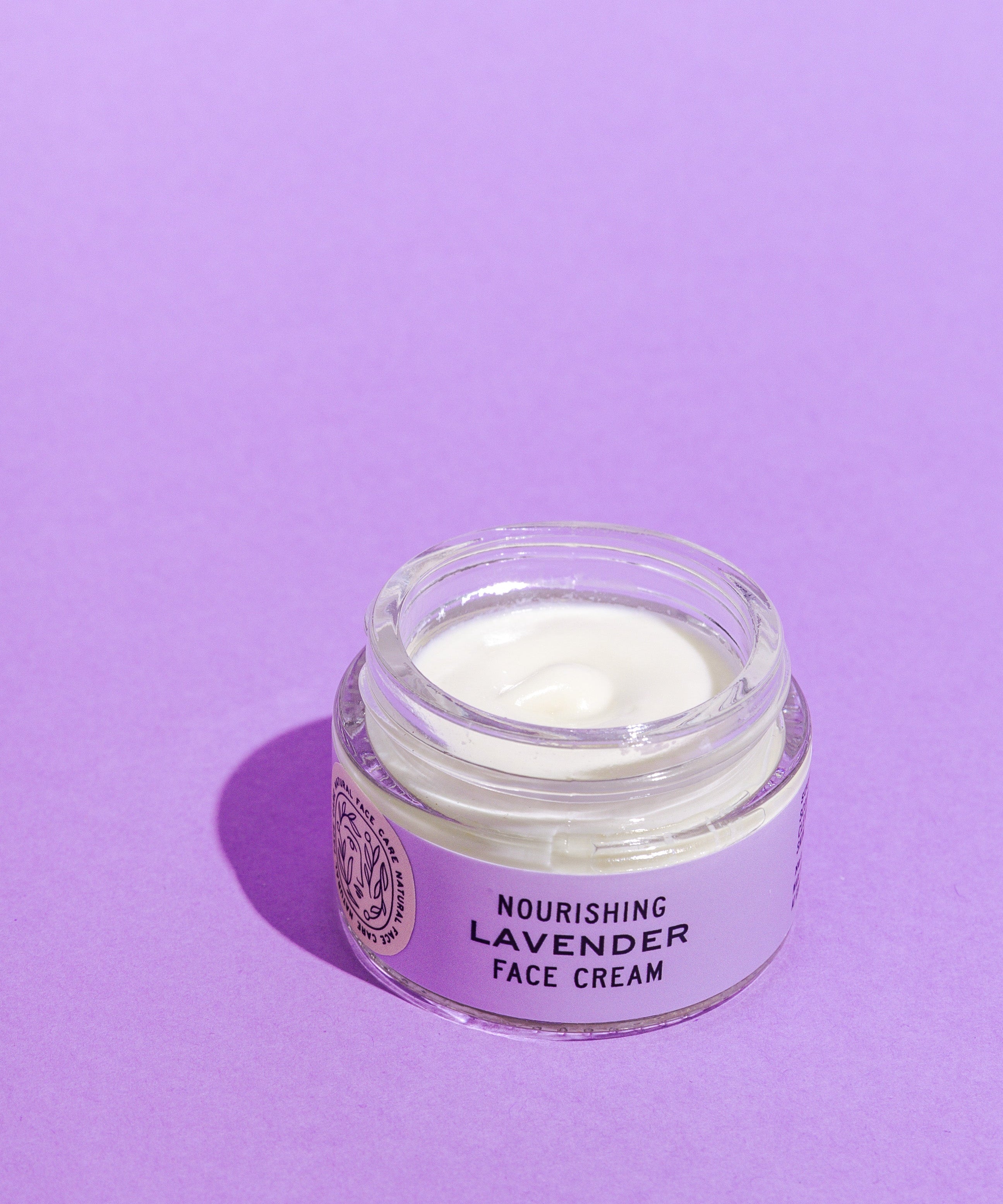 A glass jar of Good Flower Farm’s Lavender Face Cream sits open on a purple surface, revealing the creamy white, calendula-infused moisturizer inside.