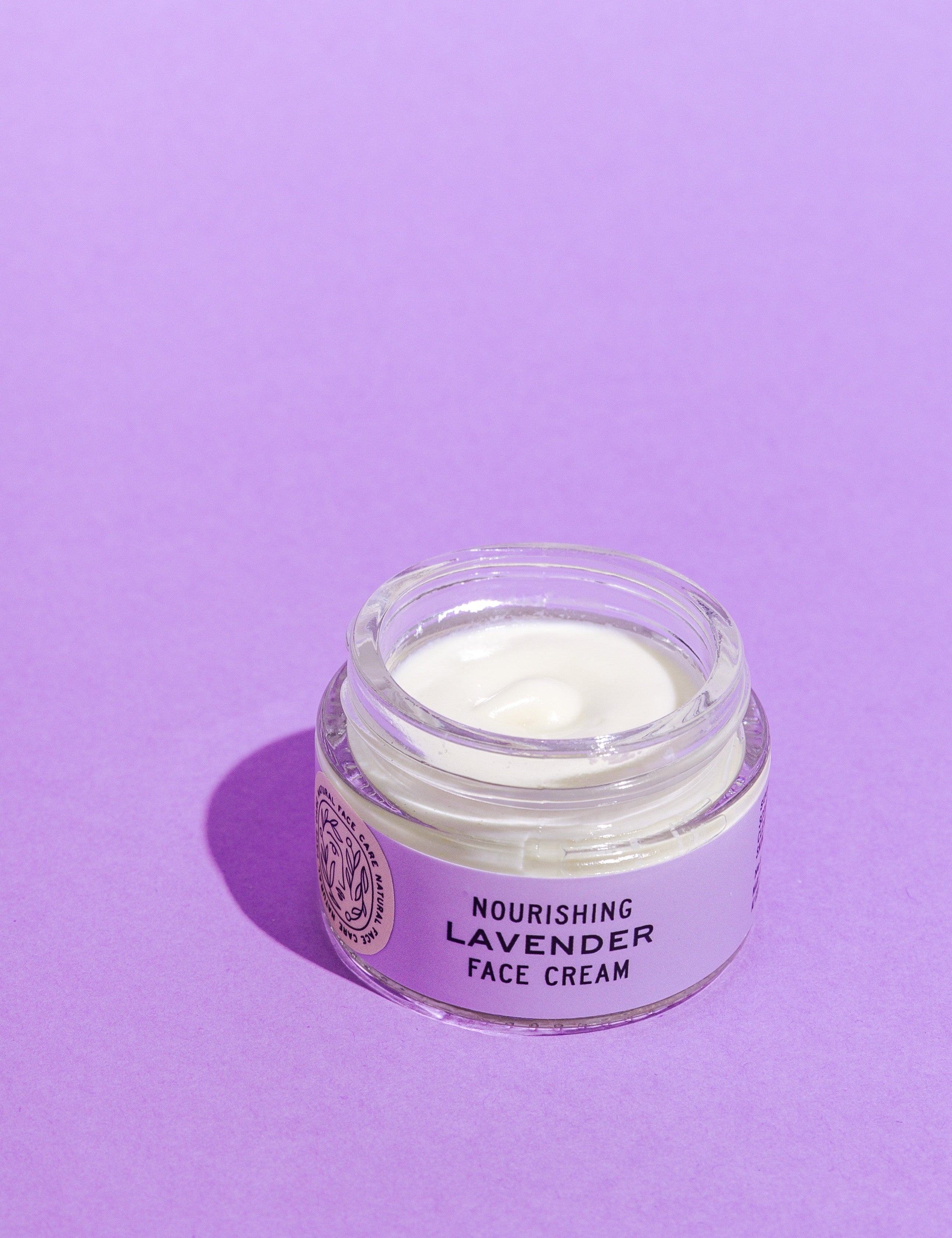 A glass jar of Good Flower Farm’s Lavender Face Cream sits open on a purple surface, revealing the creamy white, calendula-infused moisturizer inside.