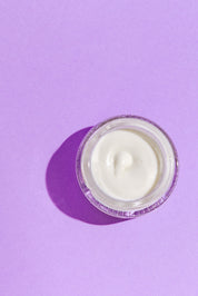 A glass jar of Good Flower Farm’s Lavender Face Cream is seen from above on a solid light purple background, with its shadow cast to the left.
