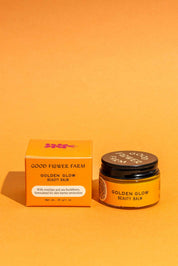 A jar and box of Good Flower Farm Golden Glow Beauty Balm, a skin repair balm, are shown on an orange background. The orange packaging features white text and purple flower graphics.