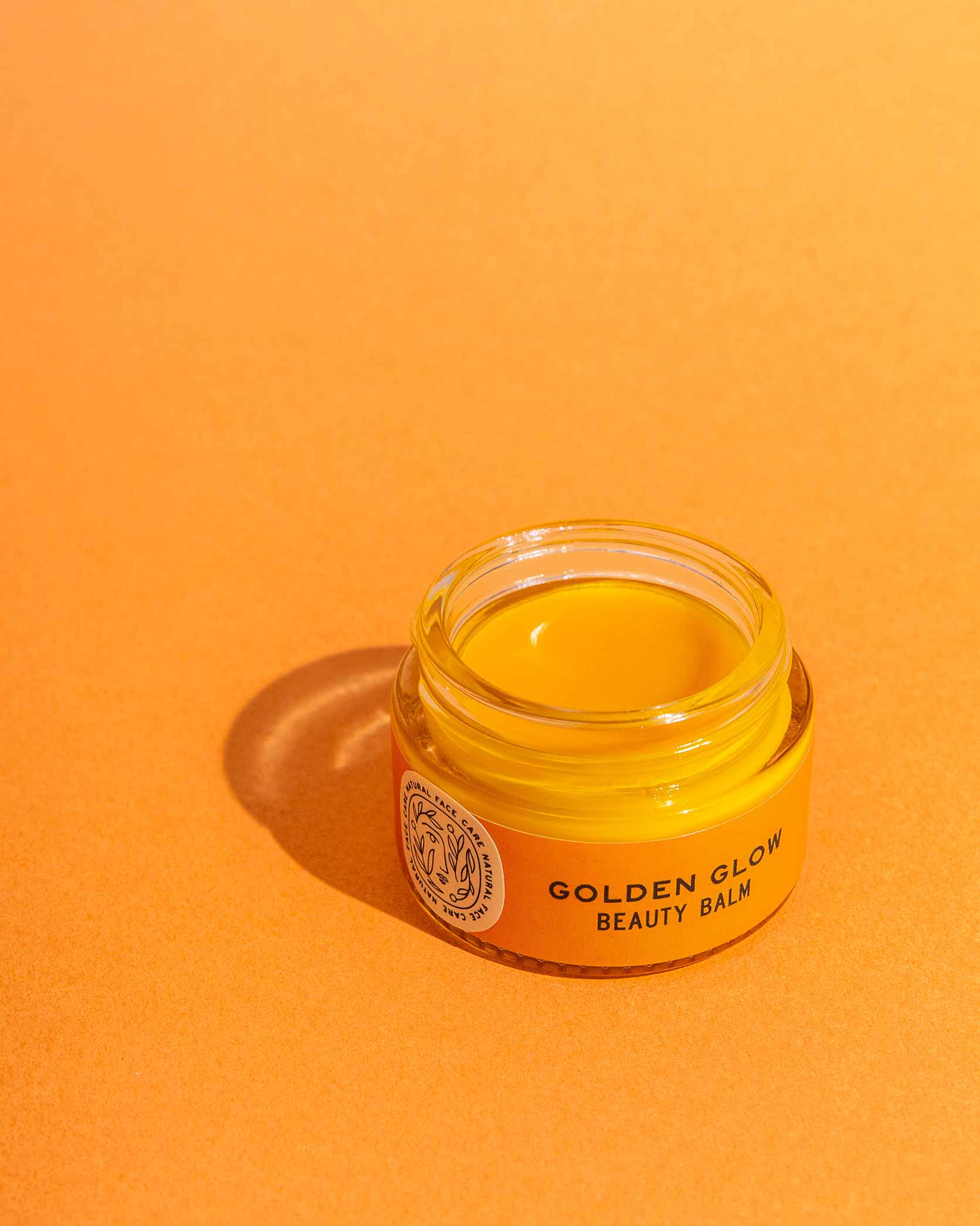 An open jar of Good Flower Farms Golden Glow Beauty Balm rests on an orange surface, its label and rich, nourishing contents illuminated by bright light—perfectly highlighting this skin repair balm.