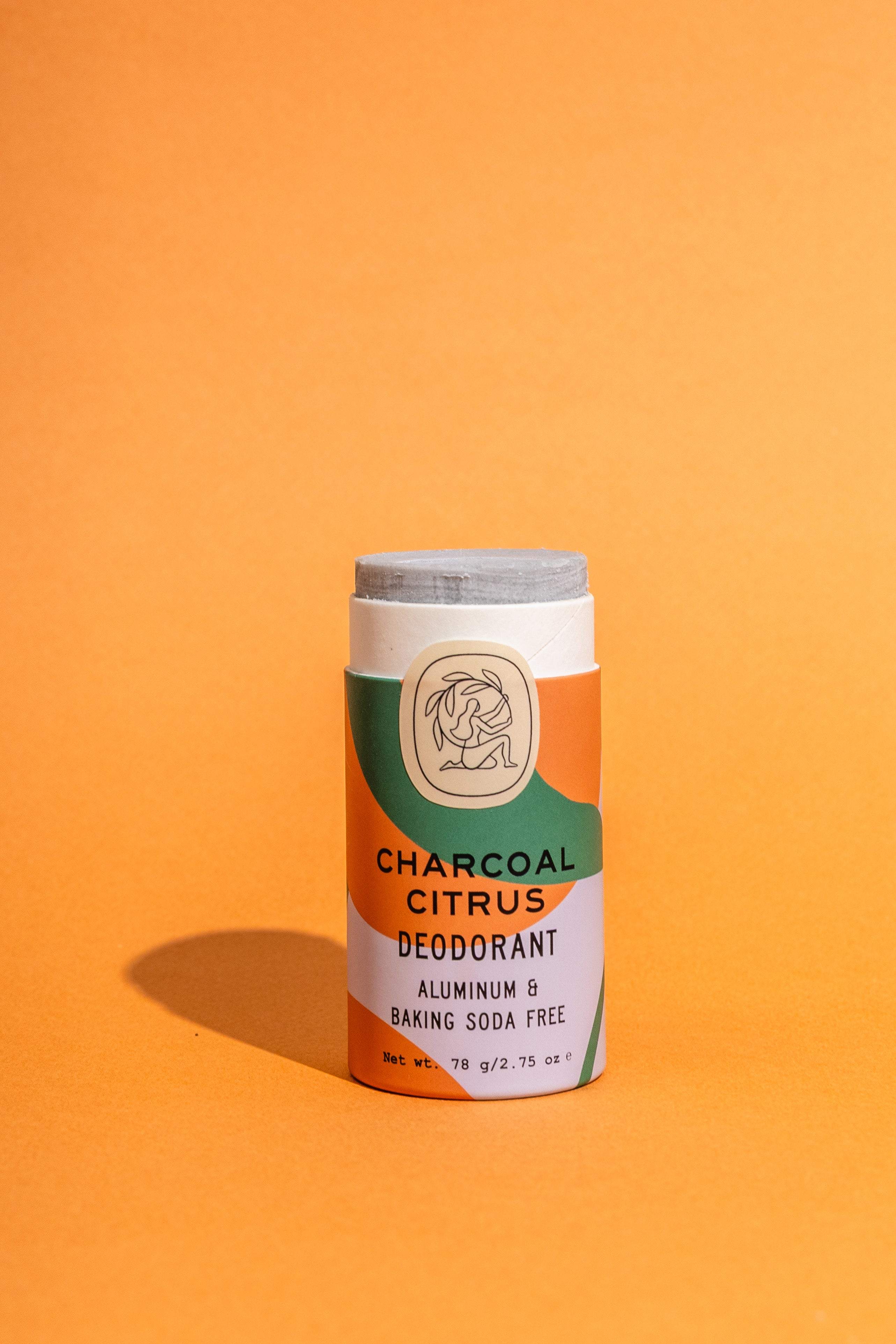 A Charcoal Citrus Deodorant Plastic-Free Tube by Good Flower Farm stands upright on an orange background, its orange, green, and cream label highlighting essential oils, aluminum- and baking soda–free formula, and 2.75 oz net weight.