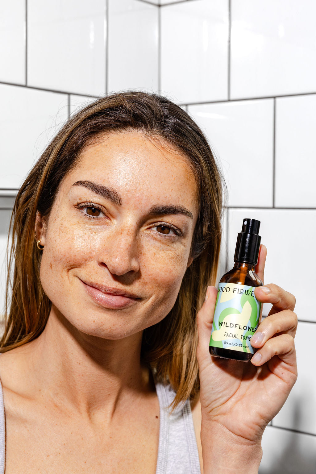 A woman with light brown hair and freckles smiles while holding up a bottle of Good Flower Farm’s Wildflower Facial Tonic in front of a white tiled bathroom wall.