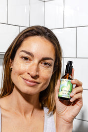 A woman with light brown hair and freckles smiles while holding up a bottle of Good Flower Farm’s Wildflower Facial Tonic in front of a white tiled bathroom wall.