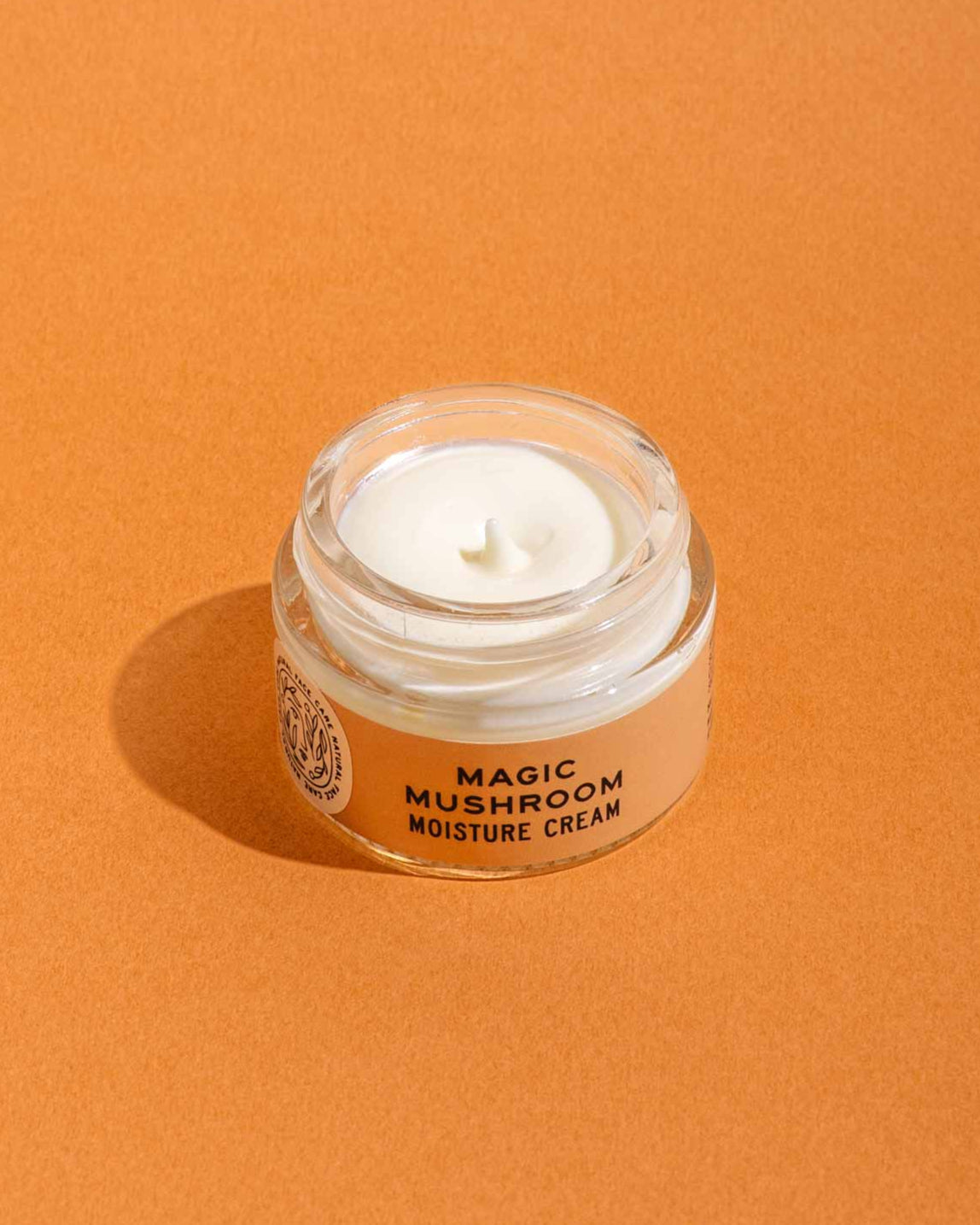 A small glass jar of Good Flower Farm’s Magic Mushroom Moisture Cream, with its lid off to display the creamy product inside, sits on an orange surface; the formula is infused with adaptogenic maitake.