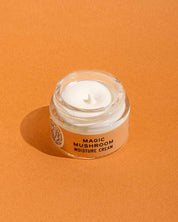A small glass jar of Good Flower Farm’s Magic Mushroom Moisture Cream, with its lid off to display the creamy product inside, sits on an orange surface; the formula is infused with adaptogenic maitake.