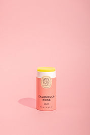 A tube of Good Flower Farm Calendula Rose Salve with a pink and cream label stands upright on a light pink background, the balm slightly exposed to showcase its soothing formula and eco-friendly packaging.