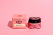 A pink jar and matching box of Good Flower Farm’s Flowers & Rose Clay Botanical Face Mask sit on a pink surface. The open jar reveals the lid, and both items are labeled for brightening and hydrating benefits.