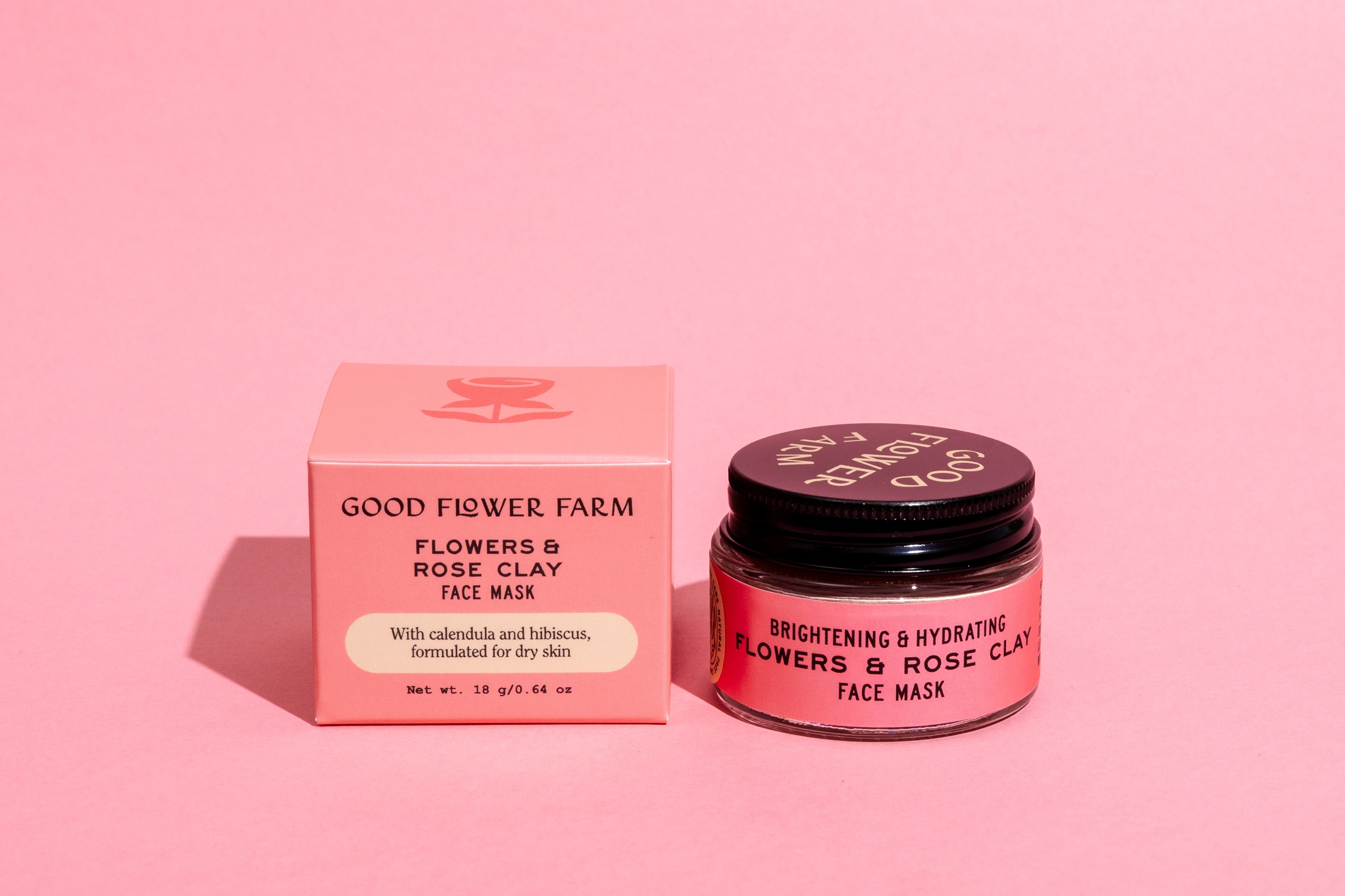 A pink jar and matching box of Good Flower Farm’s Flowers & Rose Clay Botanical Face Mask sit on a pink surface. The open jar reveals the lid, and both items are labeled for brightening and hydrating benefits.