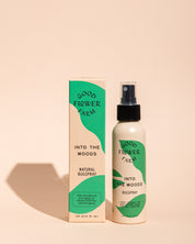 A bottle and box of Good Flower Farm Into the Woods Natural Bugspray with green and cream packaging stand upright on a neutral background, their shadows visible.