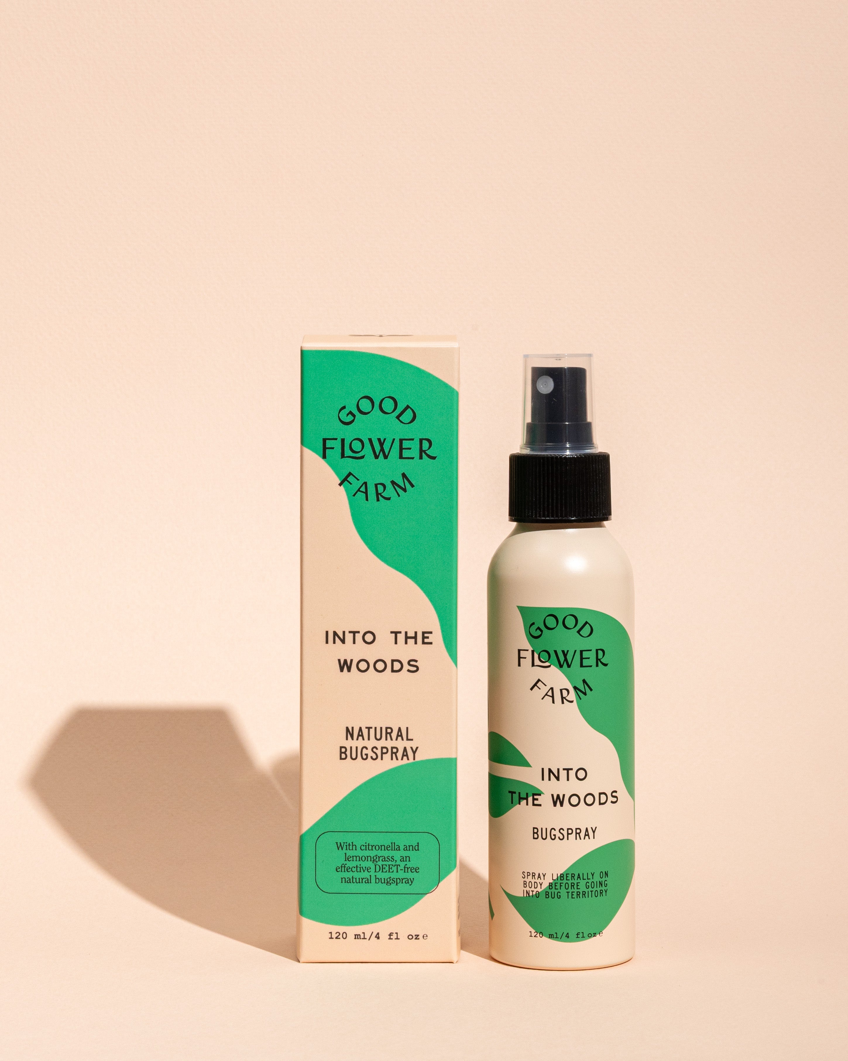 A bottle and box of Good Flower Farm Into the Woods Natural Bugspray with green and cream packaging stand upright on a neutral background, their shadows visible.