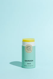 A light green tube of Good Flower Farm Garden Salve with a botanical logo and open lid reveals yellow balm inside, made from farm-grown herbs, displayed on a solid light blue background.