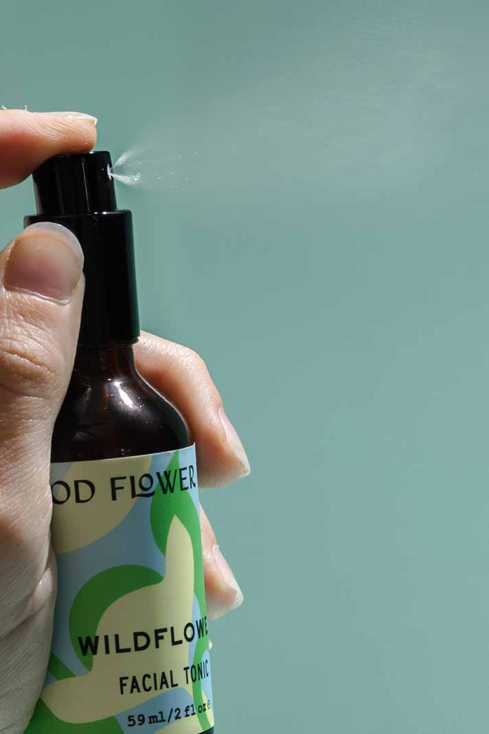A hand sprays Good Flower Farm’s Wildflower Facial Tonic, releasing a fine mist of balancing toner with wild herbal flowers against a plain, light blue background.