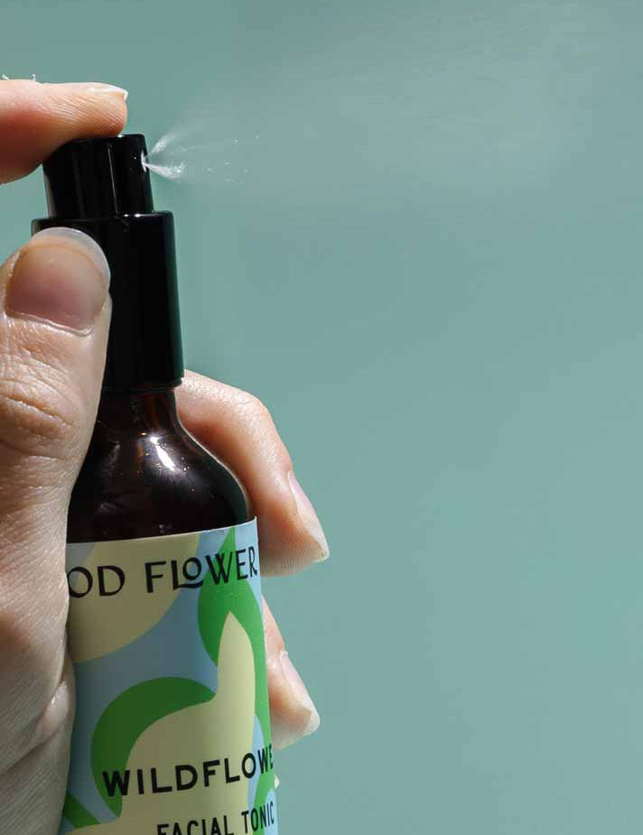 A hand sprays Good Flower Farm’s Wildflower Facial Tonic, releasing a fine mist of balancing toner with wild herbal flowers against a plain, light blue background.
