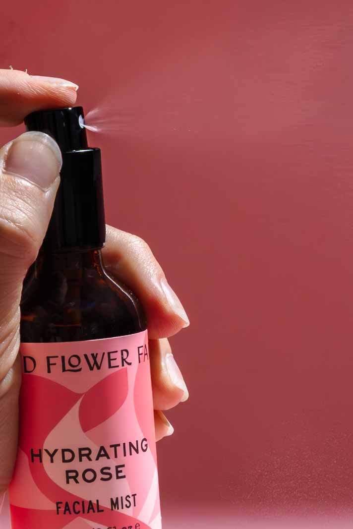 A hand sprays Good Flower Farms Hydrating Rose Facial Mist, featuring rose hydrosol and aloe vera, against a pink background. A visible mist bursts from the brown bottle, highlighting a boost of hydration.