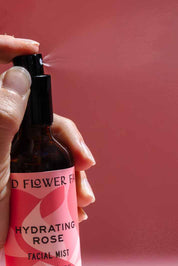 A hand sprays Good Flower Farms Hydrating Rose Facial Mist, featuring rose hydrosol and aloe vera, against a pink background. A visible mist bursts from the brown bottle, highlighting a boost of hydration.