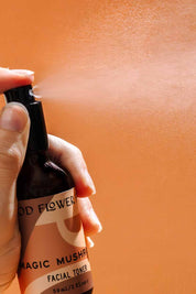 A hand sprays Good Flower Farm’s Magic Mushroom Facial Toner from a brown bottle labeled with its name, releasing a hydrating mist against an orange background.