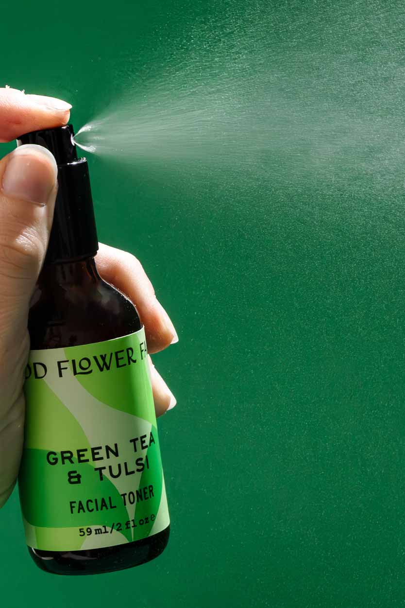 A hand sprays a mist from a brown bottle labeled Good Flower Farm Green Tea & Tulsi Facial Toner against a solid green background.
